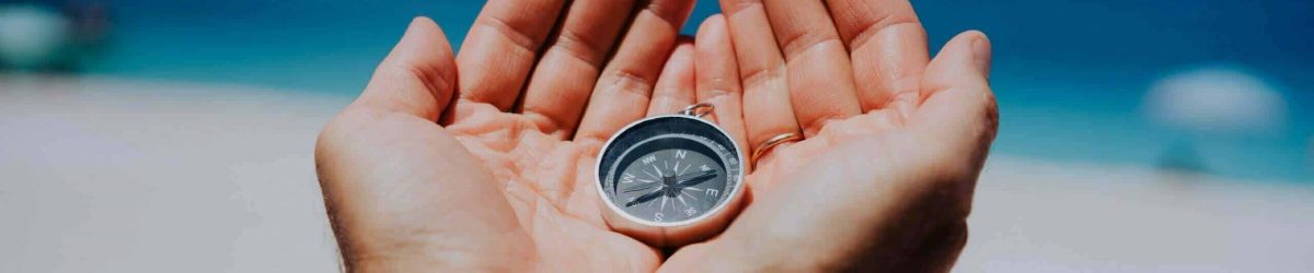 Open hand palms with metal compass on the sandy beach. Searching your way concept. Blue sea in background. Point of view pov.