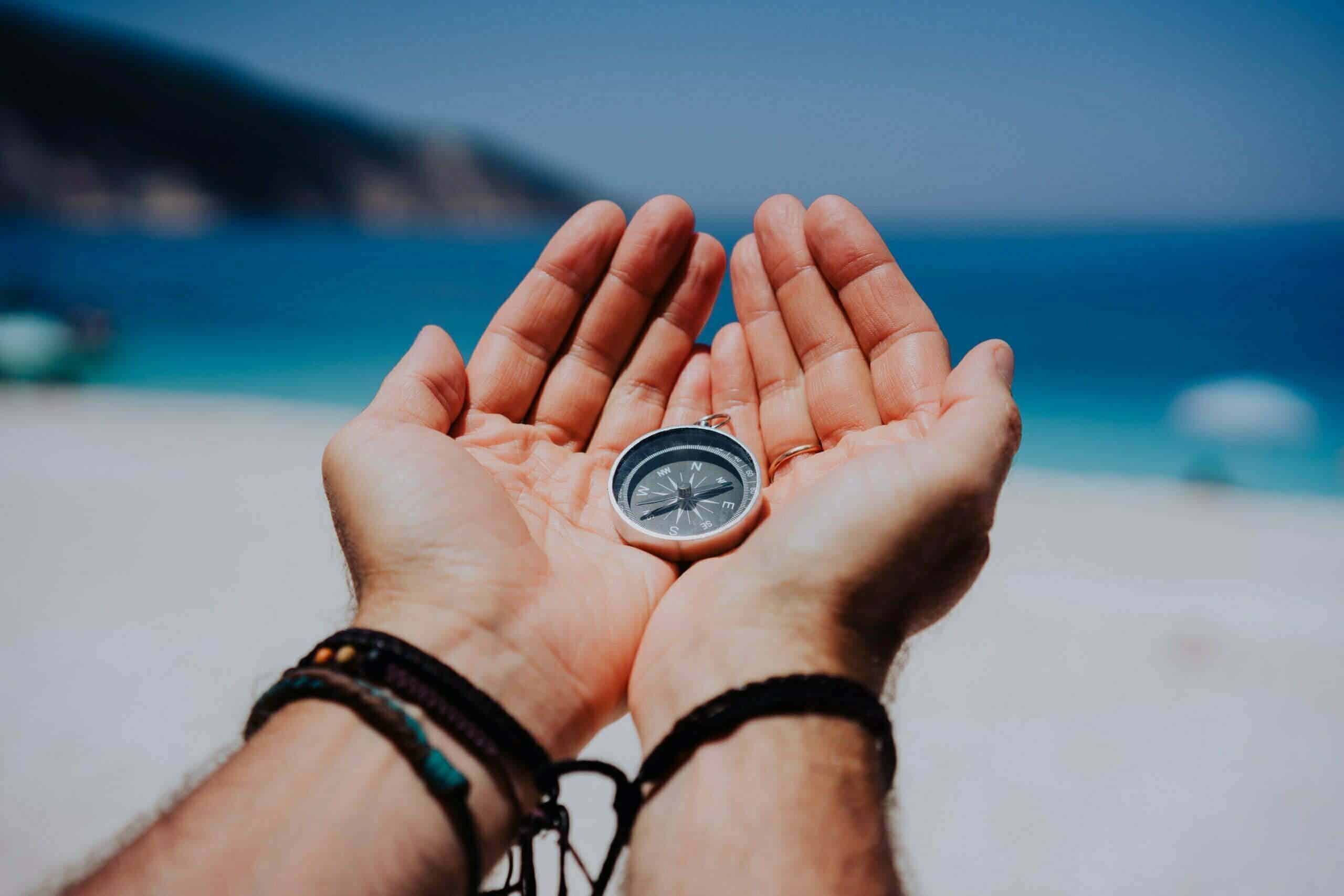 Open hand palms with metal compass on the sandy beach. Searching your way concept. Blue sea in background. Point of view pov.